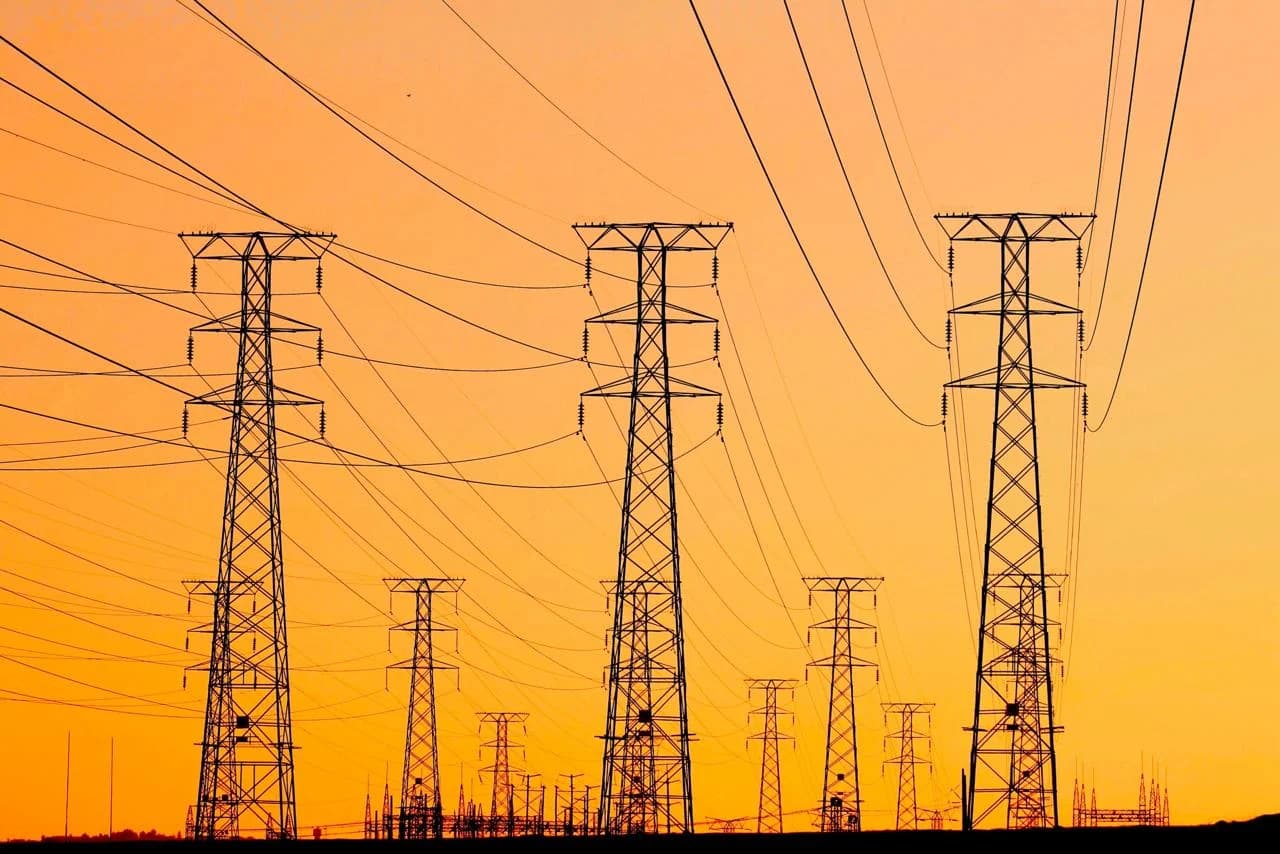 High-voltage transmission towers and power lines silhouetted against a vivid orange sky.